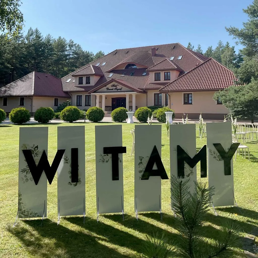 Duża tablica z czarnym napisem WITAMY ustawiona na słonecznym trawniku, w tle budynek Hotelu Tryumf otoczony zielenią i lasem.