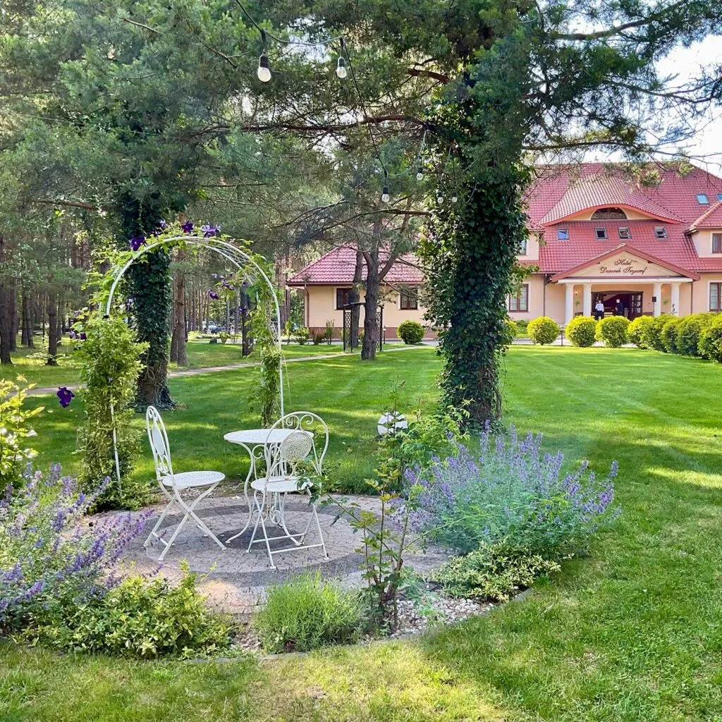 Urokliwy kącik wypoczynkowy w ogrodzie z białymi, metalowymi meblami, kwietną pergolą i rabatą z lawendą, z budynkiem Hotelu Tryumf w tle.