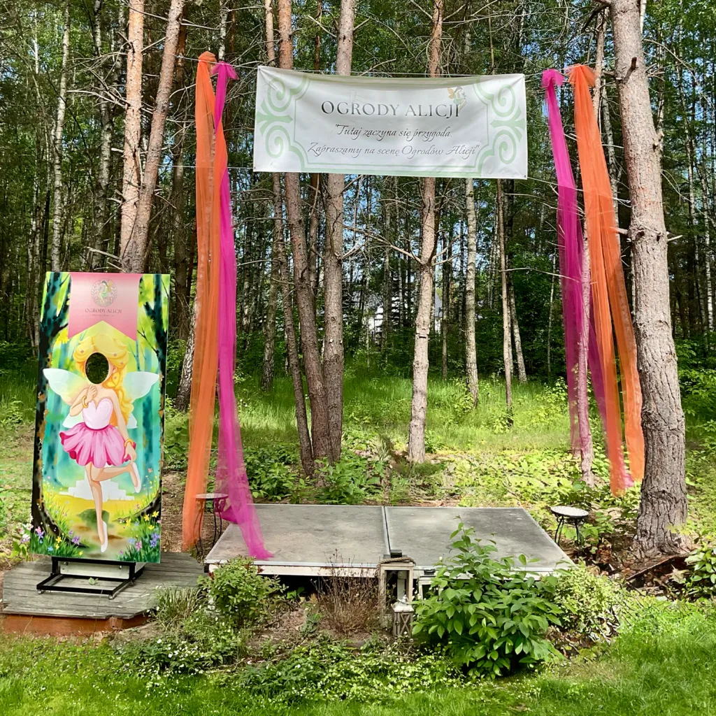 Leśna scena plenerowa ozdobiona kolorowym tiulem i banerem restauracji Ogrody Alicji, ze stojącą obok foto-ścianką z motywem wróżki do robienia zdjęć.