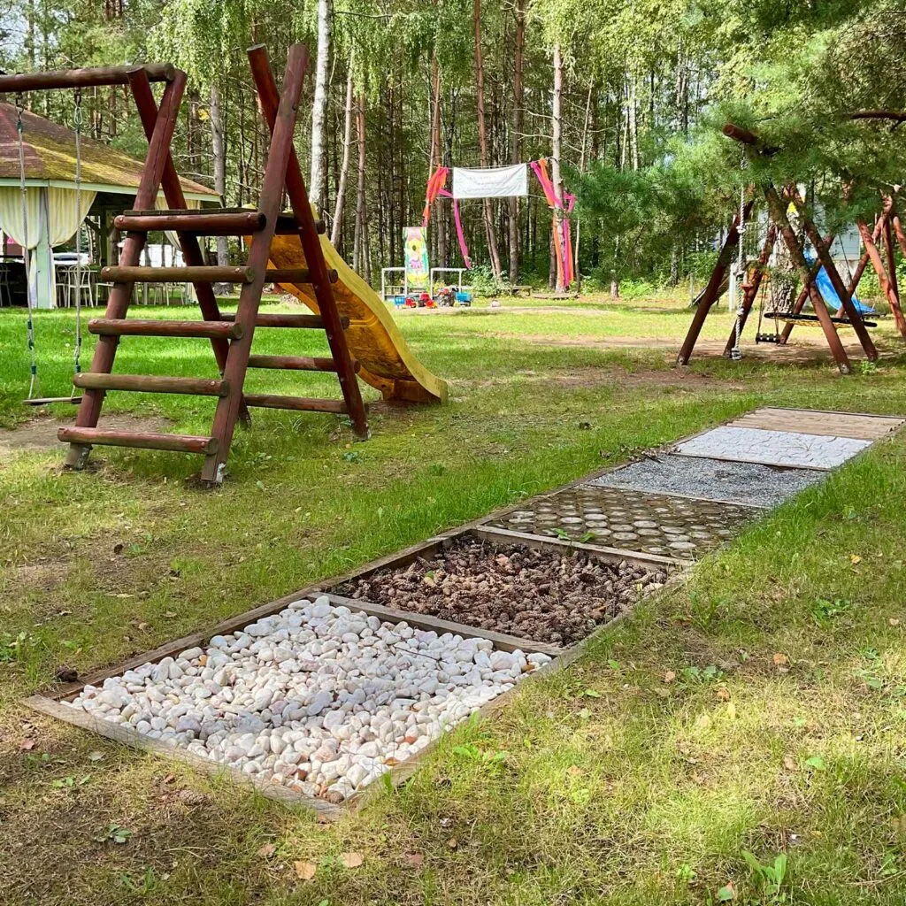 Zewnętrzny plac zabaw dla dzieci usytuowany przy lesie, ze ścieżką sensoryczną wypełnioną kamykami i szyszkami, drewnianymi huśtawkami oraz zjeżdżalnią.